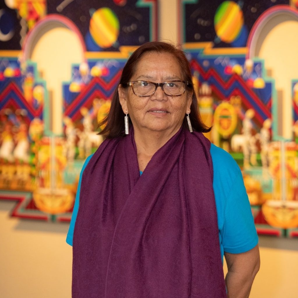 Noreen Kelly, a Navajo elder, stands before an ornate backdrop. She wears a shawl, earrings and glasses.